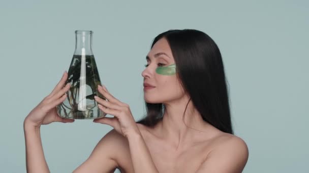彼女の目の下に緑色のヒドロゲルパッチを持つ女性は 植物のフラスコを保持しています 青い背景のスタジオにいるセミノードの女性 天然植物成分をベースとした化粧品 コンセプト — ストック動画