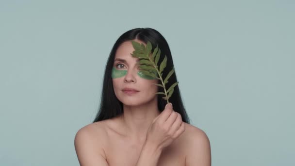 目にヒアルロンパッチを持つセミナーの女性は 顔の上に植物のスプリッグを走ります 青い背景のスタジオにいる女性が近づいている ナチュラルハーブの化粧品 美のコンセプト — ストック動画
