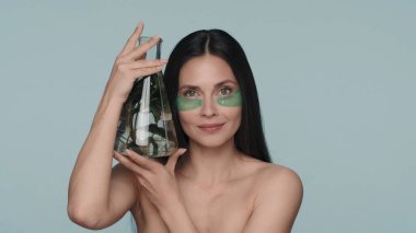 Gözlerinin altında yeşil hidrojel bantları olan bir kadın elinde bitkiyle bir matara tutuyor. Stüdyoda mavi arka planda bir kadın var. Doğal bitki bileşenlerine dayalı kozmetik ürünler. Konsept: