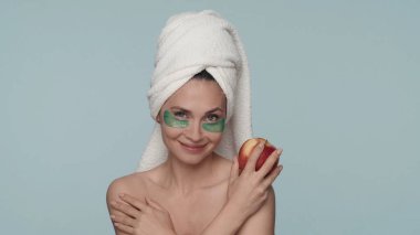 Kafasında havlu ve mavi arka planda gözlerinin altında yeşil hidrojel bantları olan bir kadın. Elinde elmayla gülümseyen bir kadının portresi. Güzellik kavramı