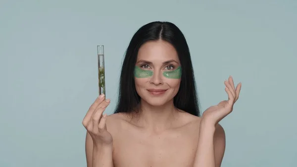 彼女の目の下に緑色のヒドロゲルパッチを持つ女性は 植物を持つテストチューブを保持しています 青い背景のスタジオにいるセミノードの女性 天然植物成分をベースとした化粧品 コンセプト — ストック写真