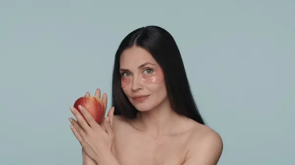 彼女の目の下にヒドロゲル赤いパッチを持つ女性は 彼女の手に熟したりんごを持っています 青い背景のスタジオのセミノードブルネットの女性が閉じています 美のコンセプト 若返り — ストック写真