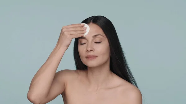 女性はスタジオで青い背景で肌の世話をする セミナーの女性は綿パッドの助けを借りて彼女の顔の皮を拭きます スキンケアのコンセプト — ストック写真