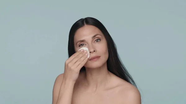 女性はスタジオで青い背景で肌の世話をする セミナーの女性は綿パッドの助けを借りて彼女の顔の皮を拭きます スキンケアのコンセプト — ストック写真