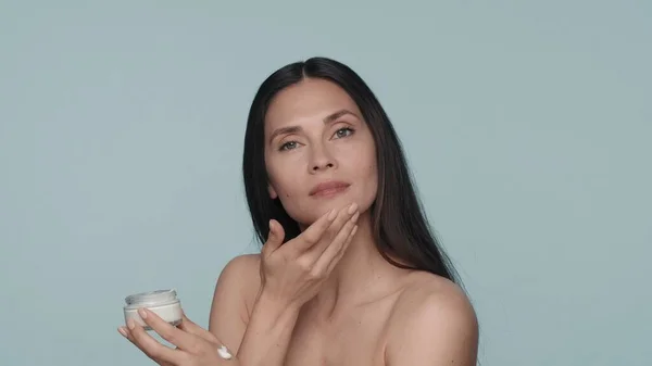 スキンケア用クリーム ブルネットの女性が顔にクリームを塗ります 青い背景のスタジオでクリームの瓶を手に入れた女性 化粧品 肌のコンセプト — ストック写真