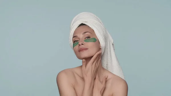 一个头戴毛巾 眼皮底下有绿色水胶斑斑的女人的画像 一个女人在蓝色背景的工作室里享受着她皮肤的纯洁和光滑 — 图库照片