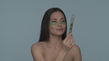 Gözlerinin altında yeşil hidrojel bantları olan bir kadın bir bitkiyle test tüpü tutuyor. Stüdyoda mavi arka planda bir kadın var. Doğal bitki bileşenlerine dayalı kozmetik ürünler. Konsept