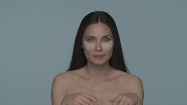 ブルネットの女性は 顔のスキンケア手順を行っています 女性は彼女の目の下のヒアルロンパッチに触れ 青い背景でそれらを滑らかにします 若返り 化粧品のコンセプト — ストック動画