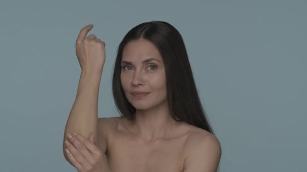 スタジオのセミノードの女性が青い背景に閉じ込められています 女性がこすり マッサージし 肘エリアの腕にローションクリームを塗ります 乾燥した肌 皮膚科 衛生学の概念 — ストック動画