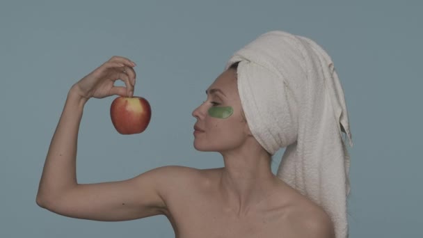 头戴毛巾 眼睛下有绿色水凝胶片 蓝色背景的女人显得很端庄 一个女人手里拿着一个成熟的红苹果 看着它 美容美发的概念 — 图库视频影像