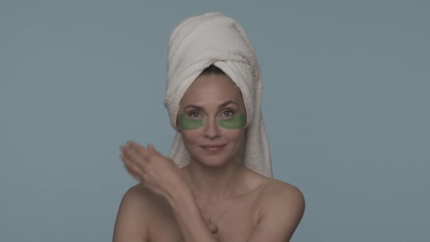 女性は目の下の皮膚からヒドロゲルパッチを取り除きます 青い背景のスタジオで彼女の頭の上にタオルを持つ女性の肖像画 美のコンセプト 若返り — ストック動画