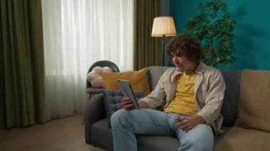Oturma odasındaki kanepede elinde tabletle günlük kıyafetlerle oturan genç bir erkek mankenin yakın plan portresi. Bir adamla görüntülü konuşma. Çevrimiçi iletişim kavramı.