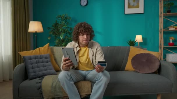 客厅里一个年轻男模的画像 男人坐在沙发上 拿着平板电脑 在应用程序中键入了一些银行卡 网上购物或汇款概念 — 图库视频影像