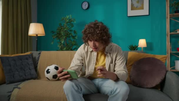 スマートフォンでサッカーの試合を見ているソファーに座っている若者の肖像画 男はその結果を待って指を横切った 彼は幸せそうに叫んでいる 彼の周りを飛んでいるお金の請求書 スローモーション — ストック動画