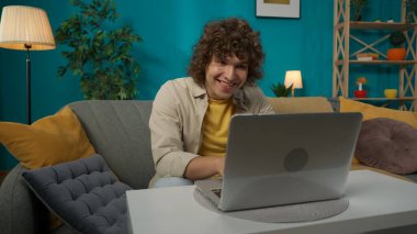 Oturma odasındaki kanepede dizüstü bilgisayarlı, günlük kıyafetlerle oturan genç bir erkek modelin yakın plan portresi. Adam bir şeyler yazıyor, internette sohbet ediyor..