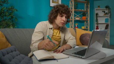 Oturma odasında günlük giysiler içinde genç bir erkek modelin yakın plan portresi. Odaklanmış yüz ifadesi olan bir adam dizüstü bilgisayarına bir şey yazıyor, deftere bir şeyler yazıyor..
