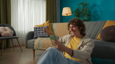 Oturma odasında günlük giysiler içinde mutlu genç bir erkek modelin yakın plan portresi. Adam yerde oturuyor ve akıllı telefonuyla komik bir film izliyor. Evde boş zamanların tadını çıkarıyorum..