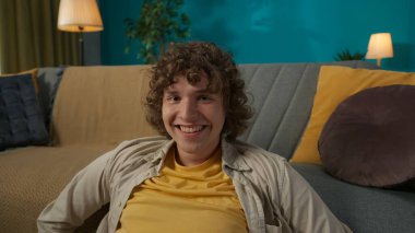 Oturma odasında yerde oturmuş kameraya bakan gündelik giysiler içindeki genç bir erkek modelin yakın plan fotoğrafı. Mutlu ve rahat gülümsüyor..