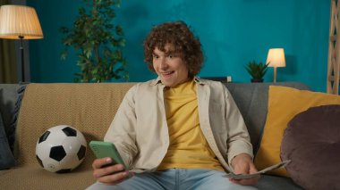 Oturma odasındaki kanepede oturan mutlu genç bir adamın portresi, akıllı telefonuyla bir futbol maçı izliyor ve elinde para tutuyor. İnsanlar mutlu ve boş vakitlerin tadını çıkarıyorlar..