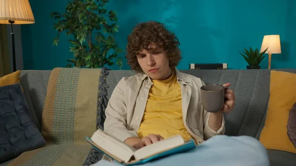 一个穿着休闲装的年轻男模坐在客厅沙发上看书 喝着杯子里的茶的画像 在家的闲暇时间 — 图库照片