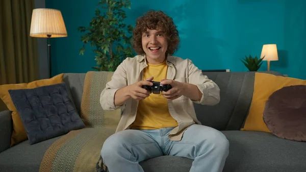 一个年轻男子坐在客厅沙发上的画像 男人用操纵杆玩游戏机 他笑得很开心 — 图库照片
