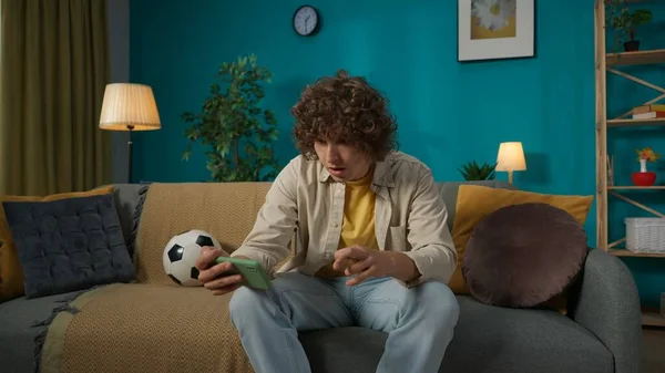 一个年轻人坐在沙发上用智能手机看足球比赛的画像 人们交叉着双手等待着比赛的结果 进行网上体育赌局 — 图库照片