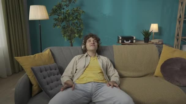 一个穿着休闲装的年轻男模在客厅沙发上放松的合影 头戴耳机的男人坐在沙发上 听着音乐 快乐地歌唱着 Hdr Bt2020 Hlg Material — 图库视频影像