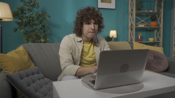 一个穿着休闲装的年轻男模坐在客厅的沙发上 桌上放着笔记本电脑 男人在打字 上网聊天 社交媒体 — 图库视频影像