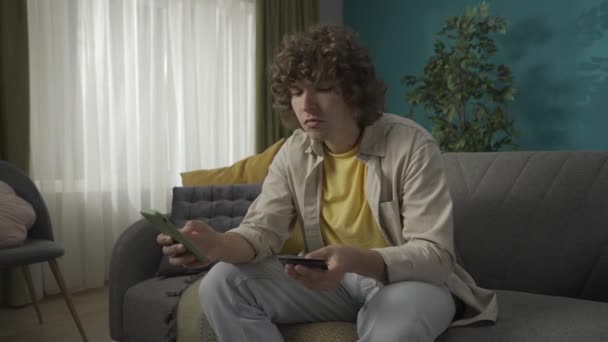 客厅里一个年轻男模的画像 男人坐在沙发上 手里拿着智能手机和信用卡 他正在应用中输入一个数字 网上购物广告概念 — 图库视频影像