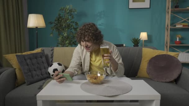 一个年轻人坐在客厅沙发上 用智能手机看足球比赛 吃小吃的画像 男人喝啤酒 享受游戏 慢动作Hdr Bt2020 Hlg材料 — 图库视频影像