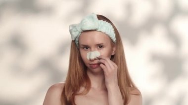 Bir kadın stüdyoda gri bir arkaplanda göz kamaştırıcı bir şekilde kozmetik işlemler yapıyor. Kafasında saç bandı olan bir kadın burnundaki siyah noktayı çıkarıp parmaklarını temiz ve pürüzsüz bir şekilde eziyor.