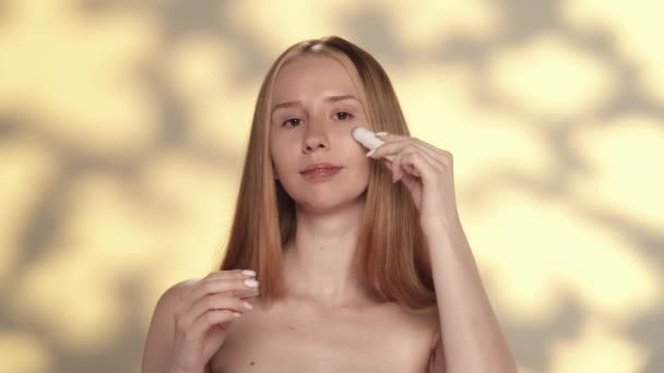 若い女性はしわを防ぐために目の周りの皮のために持ち上がるローラーのゲルを使用します ハイライト付きの黄色の背景にあるスタジオのセミノードの女性 ケアのコンセプト — ストック動画