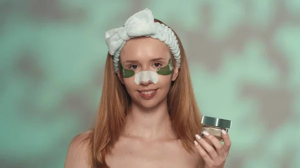 一个做化妆品的女人手里拿着一个装有奶油的罐子一个头上有发带 眼睛下面有斑斑 鼻子上有黑头石膏 绿色背景的女人 — 图库照片