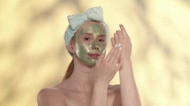 Yüzünde yeşil bir kozmetik maske olan ve stüdyoda sarı arka planda saç bandı olan bir kadın. Cildine bakarken el kremi süren bir kadın. Güzellik kavramı