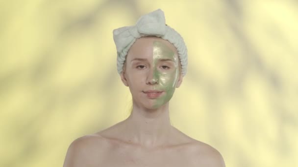 女性は顔に緑色の化粧マスクを塗ります ハイライト付きの黄色の背景にスタジオでヘアバンドを持つセミノードの女性 美容スキンケアと治療 Hdr Bt2020 Hlg材料 — ストック動画