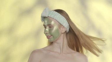Yüzünde yeşil bir kozmetik maske olan ve stüdyoda sarı arka planda saç bandı takan bir kadın. Kadın yüzünü rüzgara maruz bırakıyor. Güzellik, kozmetoloji, özen kavramı
