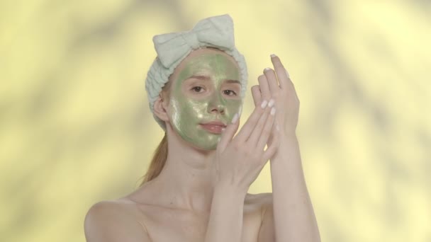 彼女の顔に緑色の化粧マスクとハイライト付きの黄色の背景にあるスタジオのヘアバンドを持つ女性 肌をケアしながらハンドクリームを塗る女性 美のコンセプト — ストック動画