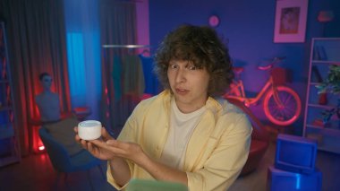 Erkek video kaydedicisi bir kavanoz yüz kremi gösteriyor, akıllı telefondan bir eleştiriyi kaydediyor. Ev stüdyosunda halka ışığının önünde video kaydeden bir adamın portresi. Kozmetik ürünleri reklamı.