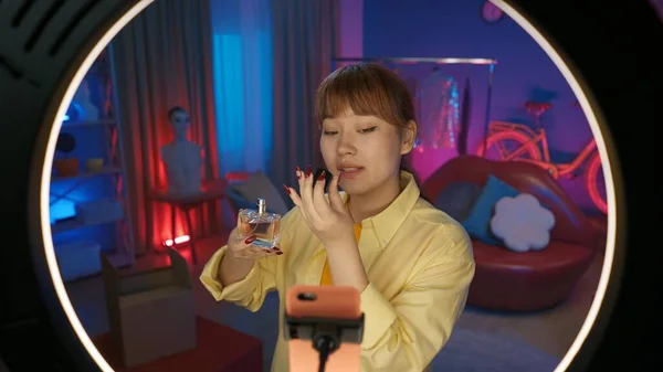 女性选美博客手拿着一瓶香水并使用它 在家里的工作室里 一个女孩在彩灯前用智能手机录下一段视频的肖像 — 图库照片