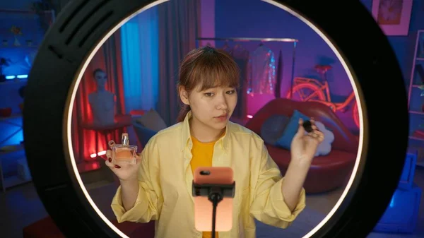 女性模特儿选美博客展示了一瓶香水 在家里的工作室里 一个女孩在彩灯前用智能手机录下一段视频的肖像 — 图库照片