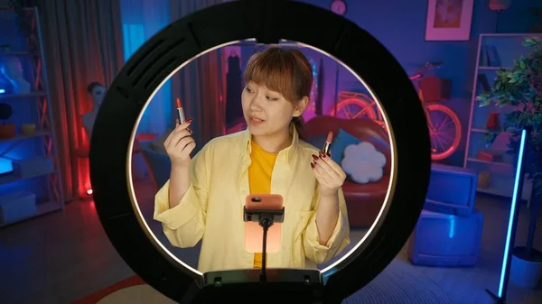 漂亮的女性博主拿着两支口红 在智能手机上拍摄 女孩在家里录音棚的光环前录制博客的肖像 美容产品概念 — 图库照片