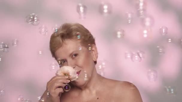 一个漂亮的金发老妇人的画像 一个中年妇女模特儿拿着一朵玫瑰花 在镜头前开心地笑着 许多肥皂泡四处飞舞 慢动作 — 图库视频影像