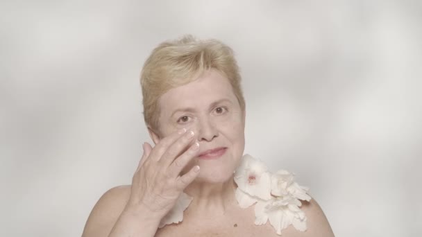 一个有魅力的老年女子的画像 一个中年妇女模特儿的近照 她用面霜把白色的角斗士花放在肩上 美丽的概念 慢动作 — 图库视频影像