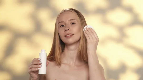 若い女性は 彼女の顔にトニック ローション ミクラー水を適用するために綿パッドを使用しています ハイライト付きの黄色い背景のスタジオに彼女の手の化粧品ボトルを持っているセミノードの女性 コンセプト — ストック写真