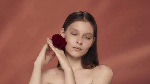 一个女人在她的脸旁边拿着一朵红玫瑰芽 摸着她柔软光滑的手的皮肤 一个有着红色背景的工作室里的优秀女人的画像 关怀的概念 — 图库视频影像