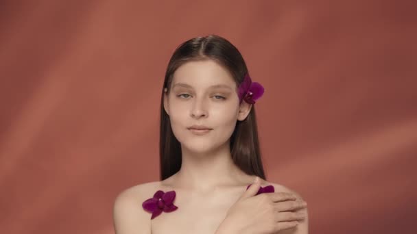彼女の髪に蘭花を咲かせた若い女性と肩に彼女の裸の肌に触れる 赤い背景にスタジオで蘭花を咲かせたセミナーの女性の肖像画 美のコンセプト — ストック動画