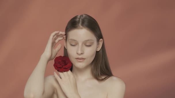 女性は顔の近くに赤いバラの芽を抱え 手の柔らかく滑らかな肌に触れます レッドバックのスタジオでセミナーの女性の肖像画 ケアのコンセプト — ストック動画