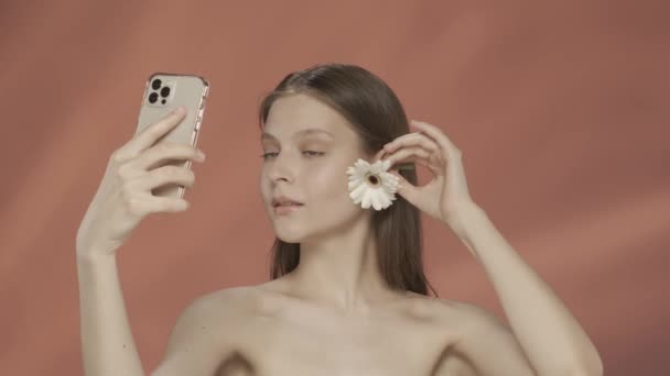 一个女人用白色的德国花瓣碰了碰她的脸 拍了一个视频 用智能手机拍了一个自拍 带着红色背景的女人站在演播室里 美容美发 美容美发和美容美发概念 — 图库视频影像
