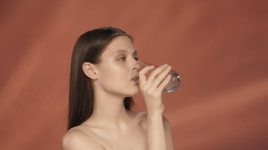 Genç bir kadın bardaktan temiz su içer. Stüdyoda kırmızı arka planda bir kadın var. Vücutta su dengesini sağlamak, sağlıklı bir yaşam tarzı. Güzellik konsepti, kozmetoloji