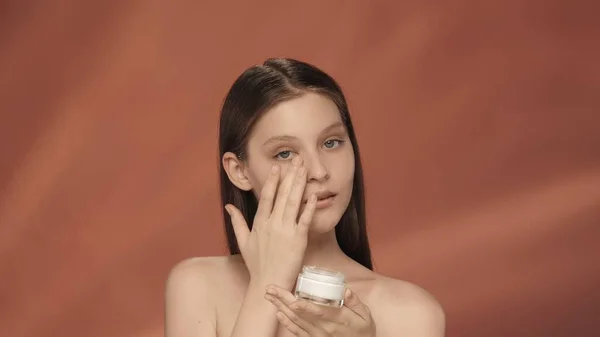 女性は化粧クリームの瓶を持ち 顔にクリームを塗ります 赤い背景のスタジオでクリームの瓶を持ったセミノードの女性 ケアのコンセプト — ストック写真